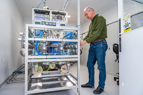 Scientist standing next to equipment encased in a metal frame with metal tubes and blue wiring