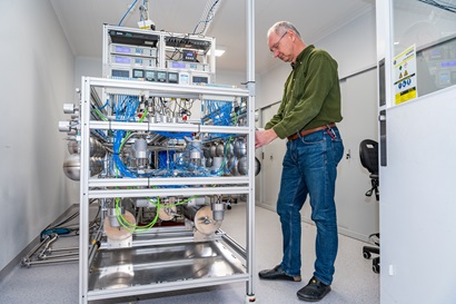 Scientist standing next to equipment encased in a metal frame with metal tubes and blue wiring