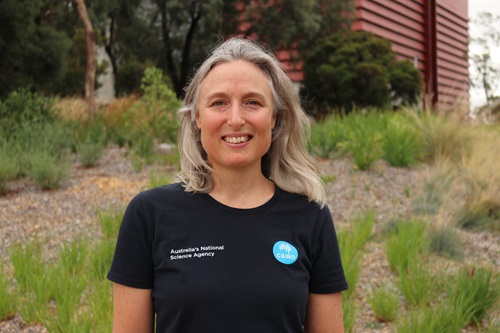 Woman in a black shirt in front of green shrubbery and building.
