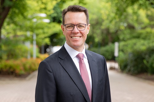 Profile photo of Kieran Perkins in a dark suit and red tie taken outside with a green trees and hedges in the background