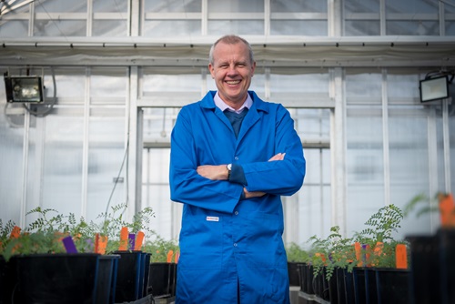 Crispin Howitt in a blue lab coat in a greenhouse between two tables of  potted plants
