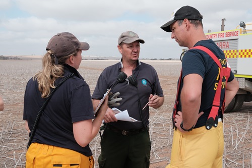 Three people in a field of stubble in discussion with a fire truck to the side