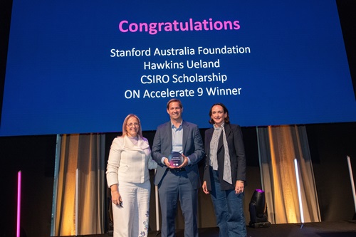 Three people on stage, award winner in the centre holding award, in front of banner which says "Congratulations. Stanford Australia Foundation, Hawkins Ueland, CSIRO Scholarship, ON Accelerate 9 winner