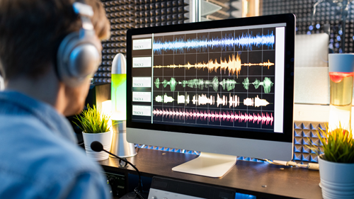 A person wearing headphones sits at a desk in a soundproofed studio, looking at a computer monitor that shows colorful waveforms across multiple audio tracks. The workspace includes a microphone, a lava lamp, and small potted plants.