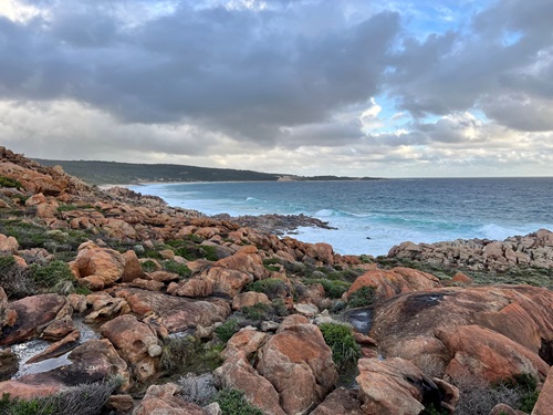 Reddish boulders and rocks leading down into the sea wash of a bay, with a cloudy sky overhead