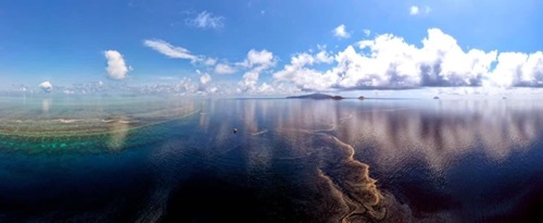 Wide angle lens view of sea surface showing the reef underwater and coral larvae plume on the surface with clouds in the background.