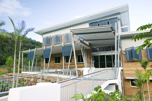Modern building with a sloped roof and blue window shades, surrounded by tropical plants and trees, located in a lush green setting.