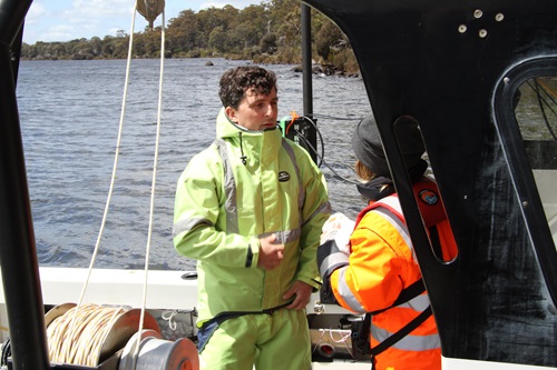 Two people wearing high-visibility waterproof gear are standing on a boat, preparing for work on a lake. One person is dressed in bright green protective clothing, while the other is in an orange jacket with reflective stripes and a dark beanie. They appear to be discussing equipment or procedures. The boat has visible ropes, a winch, and other gear, suggesting a survey or research activity. In the background, the lake stretches out with rippling water and a tree-lined shore under partly cloudy skies.