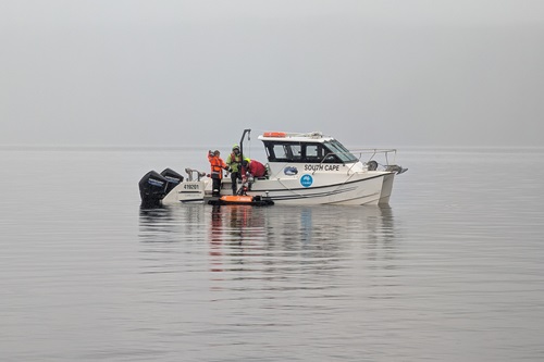 A white research vessel named South Cape floats on calm, misty waters of Lake St Clair. Two people in high-visibility gear are on the rear deck deploying an orange autonomous surface vehicle equipped with sensors.