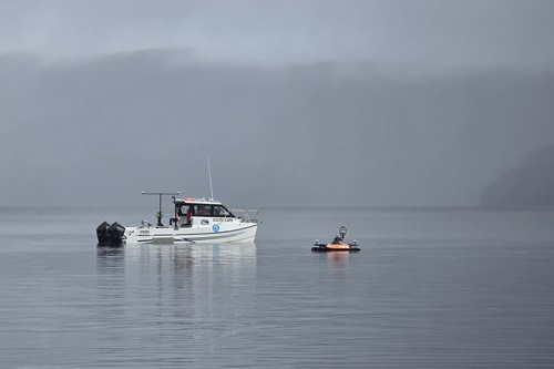 A research vessel floats on calm, mist-covered waters with a small orange autonomous surface vehicle positioned nearby. The scene is framed by a grey, foggy backdrop with faint outlines of hills in the distance.