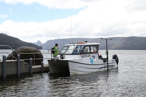 A white research vessel named South Cape is moored at a lakeside jetty, with calm water in the foreground and forested hills and mountains in the background under a partly cloudy sky.