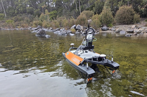 An orange and black uncrewed surface vessel floats in shallow, clear water near a rocky shoreline with dense green vegetation in the background. The vessel is equipped with sensors and technical instruments for aquatic surveys.