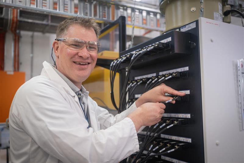 Person in a laboratory setting wearing safety glasses and a lab coat, at a work station with lithium-ion battery cells and diagnostic equipment. 