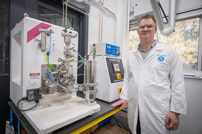 Laboratory setup featuring specialised equipment for lithium-ion battery research, including a white machine with metal components, wiring, and control panels, alongside another device with labeled buttons. A person wearing a white lab coat stands next to the equipment in a research facility.