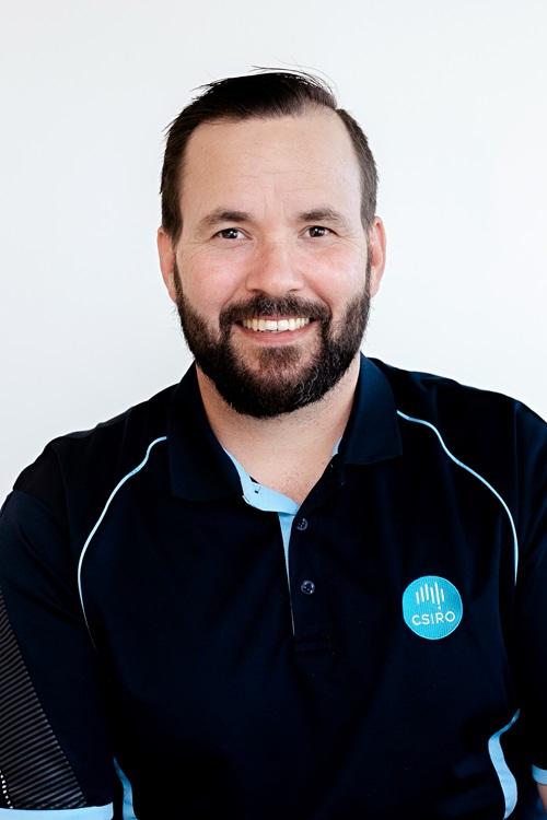 A man with short hair and a beard wearing a dark polo shirt with light blue accents and a circular logo on the chest, standing against a plain white background.