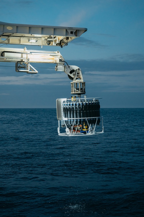 A circular piece of scientific equipment being held over the ocean at night by a boom.