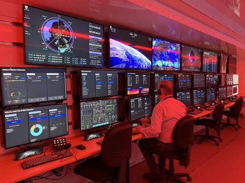 Interior of a control room with multiple monitors displaying maps, graphs, and real-time data. A person is seated at a workstation actively monitoring the screens.