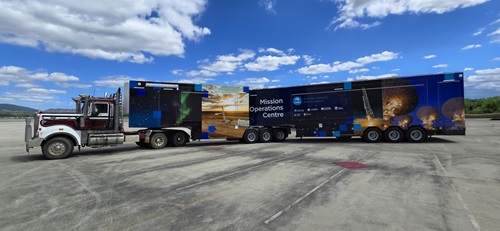 Side view of a large multi-trailer truck labeled "Mission Operations Centre," featuring external compartments and staircases, designed as a mobile command center.