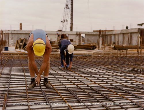Two construction workers are installing steel reinforcement bars on a concrete floor at an early-stage building site. Both are dressed in protective gear including hard hats. One worker is bent over in the foreground, positioning the rebar, while the other is working further back. The site is surrounded by construction materials and equipment, indicating active development