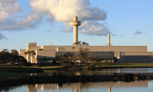 Front view of a large industrial building with a central cylindrical tower, surrounded by trees and reflected in a nearby body of water under a partly cloudy sky