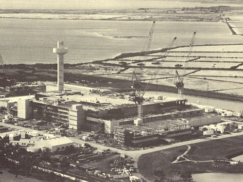 Aerial view of a large coastal construction site featuring cranes, partially built structures, and a tall central tower, surrounded by water channels and land plots