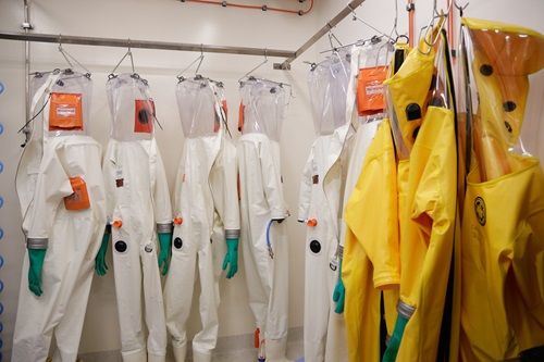Protective biohazard suits hanging in a storage area, including white suits with green gloves and transparent hoods, and yellow suits with black gloves, used for working in high-containment environments