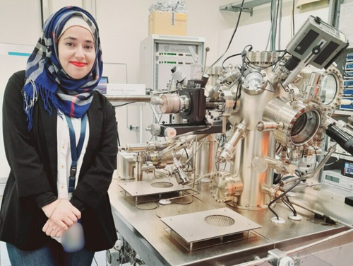 A woman in a black blazer, white shirt, and blue head scarf with red, white and grey stripes standing beside a complex scientific instrument in a laboratory.