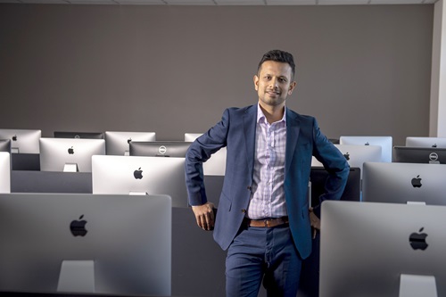 erson in a blue suit and checkered shirt standing in a computer lab with rows of computrts leaning against a desk.