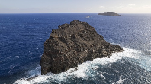 A rocky island in the ocean with a ship and another rocky island in the background.