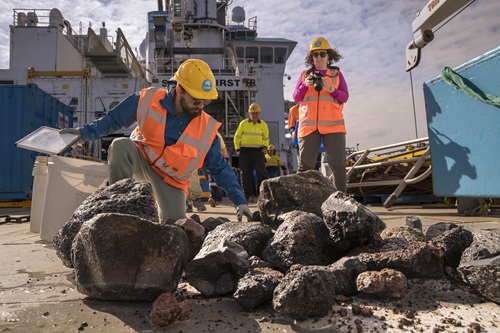 A group of people in hi vis vests and hard hats look through rocks on the back deck of a ship.