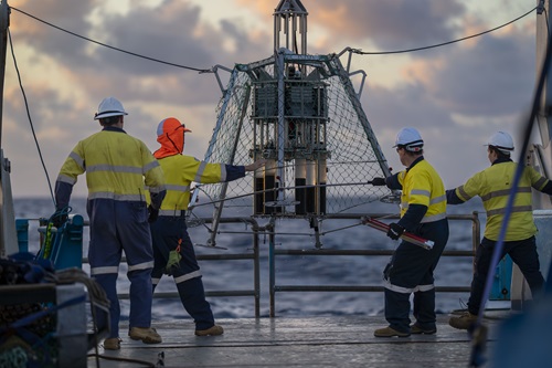 A piece of scientific equipment that looks like a lunar lander being lowered onto the deck of a ship by a group of people.