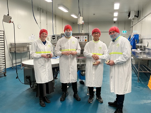 Four people in white lab coats with yellow reflective stripes and red hairnets stand in a brightly lit industrial kitchen, holding small containers of food. Two individuals are shaking hands. Stainless steel equipment, a wall clock, and machinery are visible in the background.