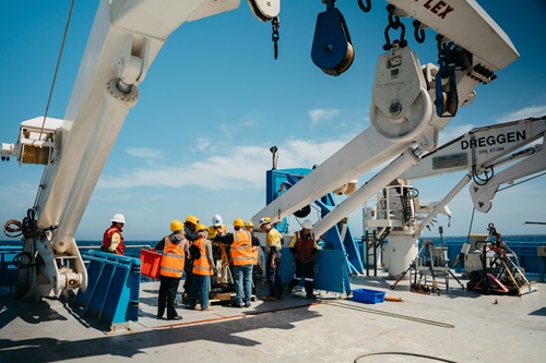 A group in safety gear gathers on the open deck of a research vessel near large cranes and winches during a scientific operations briefing at sea.