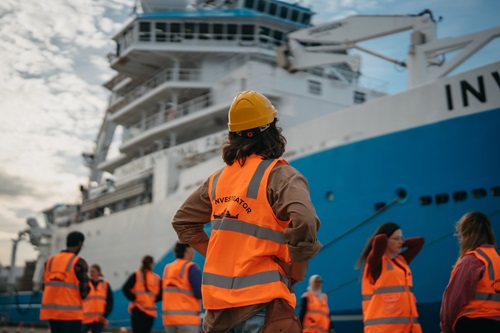 People in high-visibility vests and hard hats stand on the deck of a large blue-and-white research vessel, looking up at the ship’s superstructure.