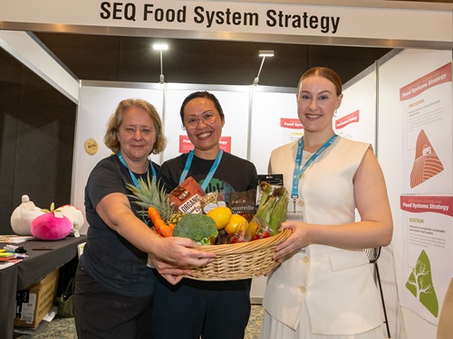Three people holding a basket of fruit and vegetables facing the camera with a conference booth behind