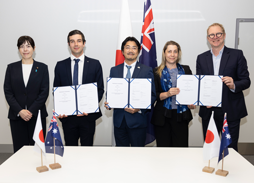 Five people standing in a row behind a table with small flags of Australia and Japan and holding copies of the signed agreement. Left to right: Japan's Minister in Charge of Economic Security Kimi Onoda, Anthony Chesman (CSIRO), Jungo Okai (Fujitsu), Petra Andren (Quantum Australia) and John Carroll (Monash University).
