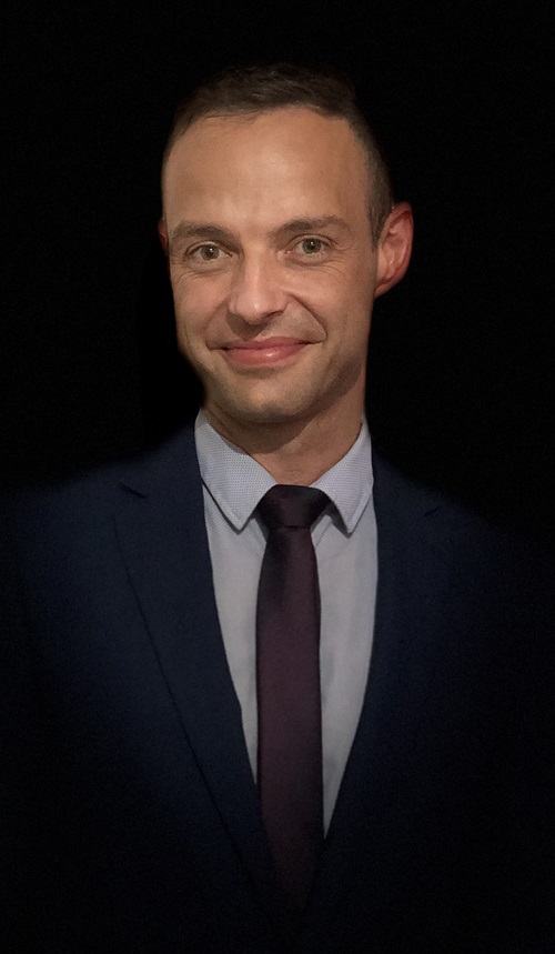 Individual dressed in a dark suit and tie with a light collared shirt, photographed against a black background.