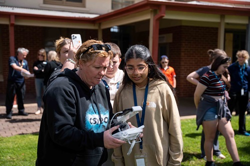 Two people standing outside on a lawn, one demonstrating how to operate a small drone controller while others gather in the background.