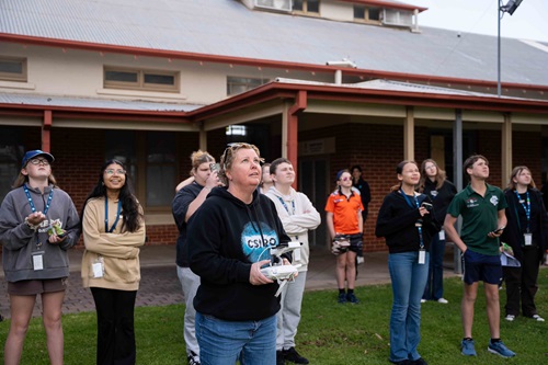 A group of people standing on a lawn outside a building, watching as a person in the foreground operates a drone controller.