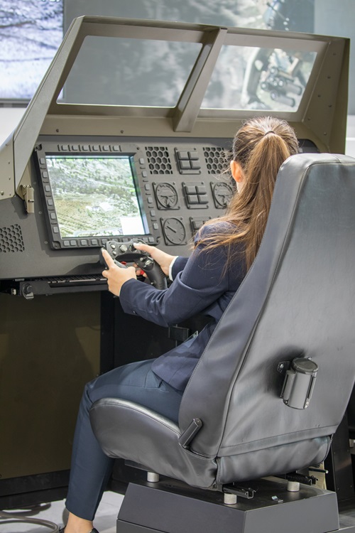 A person seated in a simulator chair operating a vehicle-control console, using hand controls and viewing a large screen displaying terrain.
