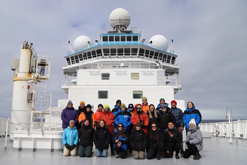A group of people wearing cold‑weather gear pose for a group photo on the bow of a research vessel, with the ship’s bridge and radar domes towering behind them under an overcast sky.