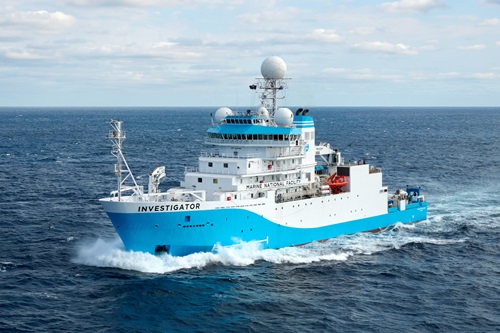 A large blue‑and‑white CSIRO research vessel named “Investigator” travels through open ocean, creating white spray as it cuts through the waves under a partly cloudy sky.