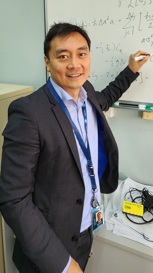 A man wearing a suit jacket and with an office lanyard around his neck smiles at the camera. He is writing on a whiteboard. 