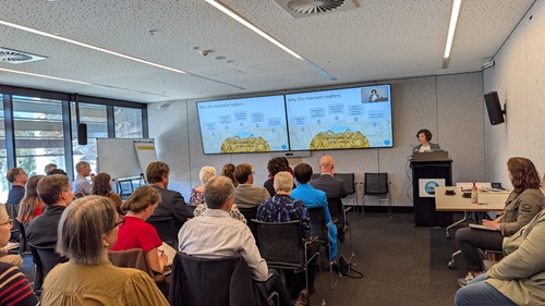 Seminar room with participants on chairs facing two screens and presenter at the front of the room,