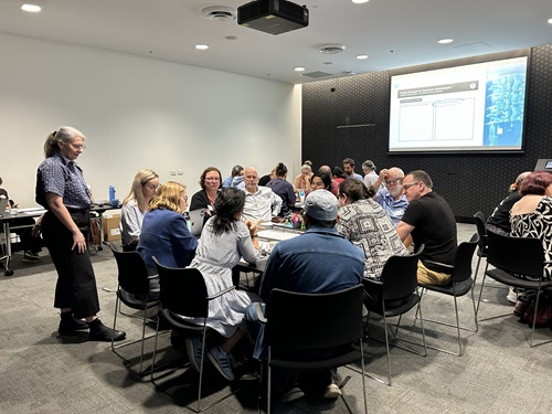 A workshop taking place in a seminar room with tables of participants and a screen at the front of the room