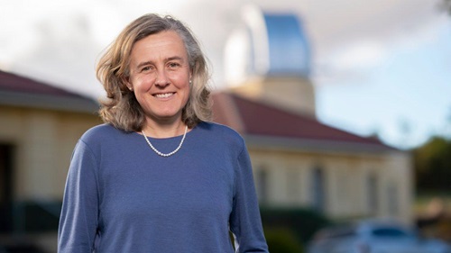 A person standing outdoors in front of a blurred building with a dome-like structure in the background. The person is centered in the frame, wearing a long-sleeved blue top and a short beaded necklace. The background includes soft-focus architectural elements and a parked vehicle, with natural daylight illuminating the scene.
