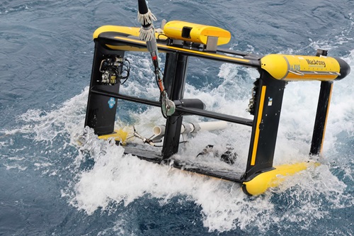 A large, yellow and black box-shaped piece of scientific equipment being towed through the ocean.