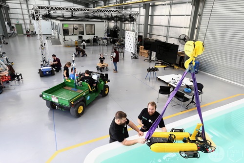 Large robotics lab with multiple researchers working on robots and equipment, including ground vehicles and robotic arms, inside a spacious industrial workshop with testing structures and tools.