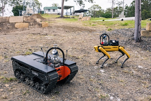 Outdoor test site with two autonomous robots: a tracked ground vehicle and a four‑legged robot, positioned on rough terrain with pipes, rocks and research infrastructure in the background.