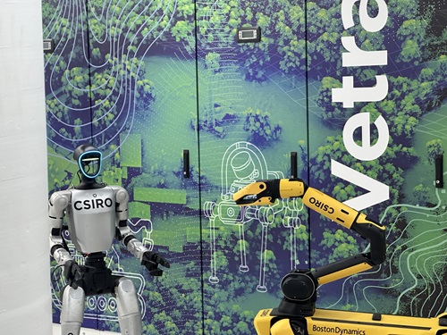 Humanoid robot with “CSIRO” branding stands next to a yellow robotic arm in front of a colourful Vetra data centre wall featuring aerial landscape graphics and large vertical “Vetra” text.